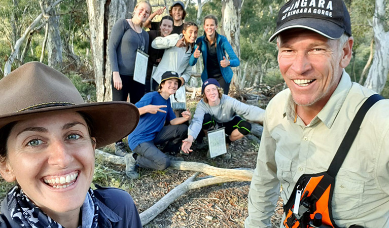 2 laughing YouthAdvance instructors, with a group of young people smiling and posing in the background in a forest setting. Photo credit: Keith McReynolds &copy; YouthAdvance