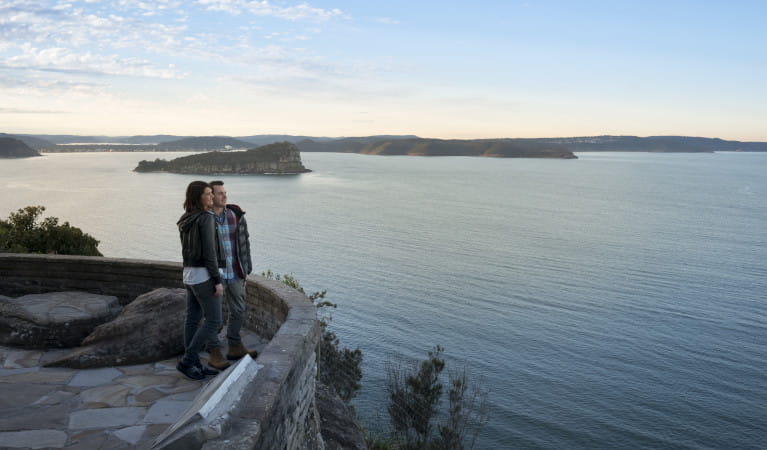 A woman and man standing at sandstone lookout, water and coastline in background. Photo: John Spencer &copy; DPIE