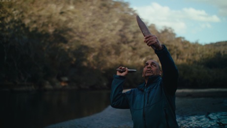 Dharawal Aboriginal Elder Dean Kelly with clapsticks by a bushland waterway in Royal National Park.  Photo: Aristo Risi/DCCEEW &copy; DCCEEW