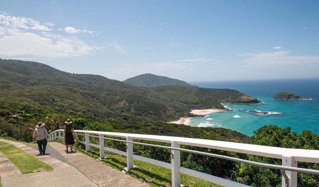A guide and visitor take the 10-minute walk back down from Smoky Cape Lighthouse after the tour, enjoying the view of sparkling ocean, beaches and islands, Hat Head National Park. Credit: Louise Walpole/DCCEEW &copy;DCCEEW