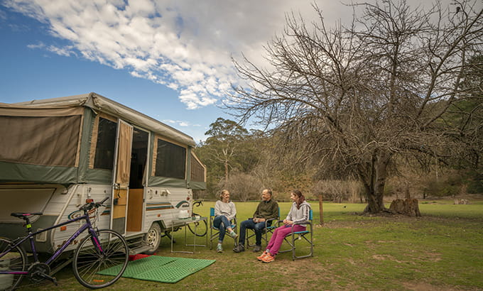 Three people sit on camp chairs beside a caravan at Wombeyan Caves campground. Photo: John Spencer/DPIE