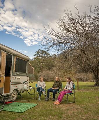 Three people sit on camp chairs beside a caravan at Wombeyan Caves campground. Photo: John Spencer/DPIE