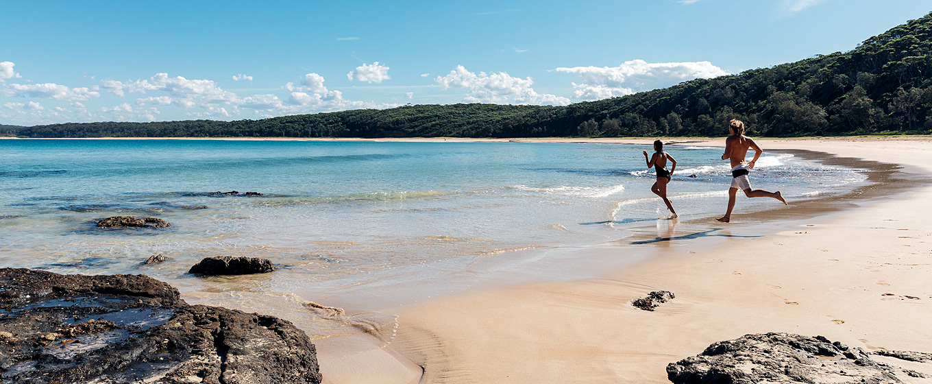 A woman and man run into the water at a beach in Murramarrang National Park. Photo: Melissa Findley