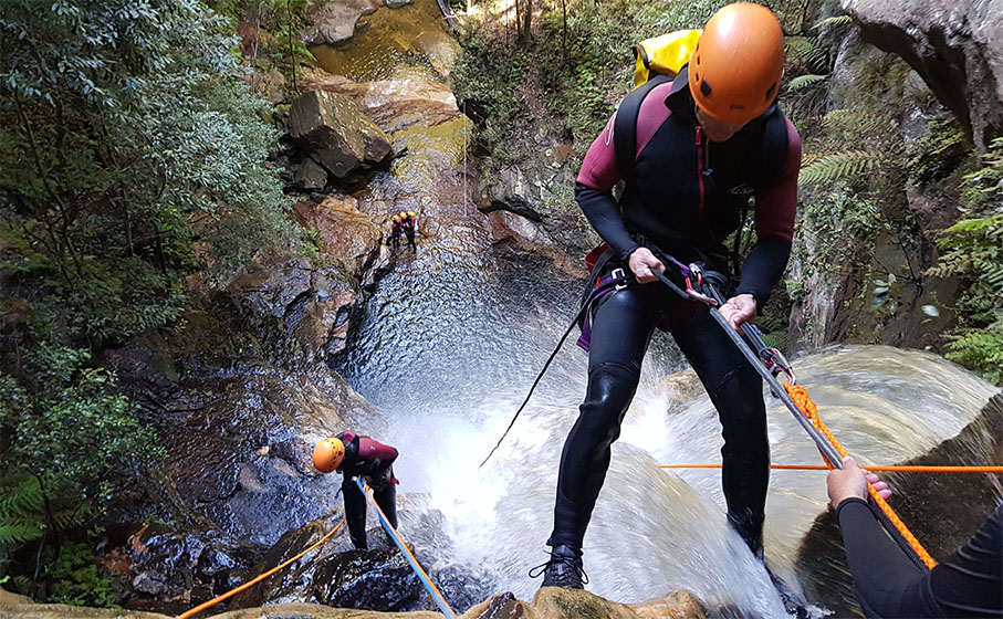 2 people in wetsuits descend on ropes past a waterfall into a steep canyon. Photo credit: James Waddell © Eagle Rock Adventures