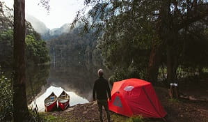 A man stands near a tent next to a river, with 2 canoes by the bank. Photo: Tim Clark/OEH