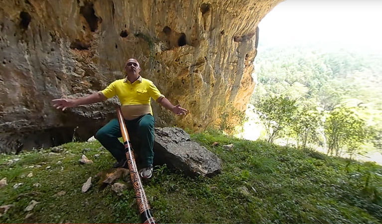 Video still of a man with a didgeridoo sitting on a rock at South Glory Cave entrance in Kosciuszko National Park. Photo: DPIE