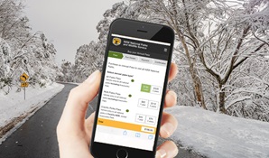 A hand holds a mobile phone in front of a snow-covered road in Kosciuszko National Park. Photo: Murray Vanderveer Artwork: DPIE