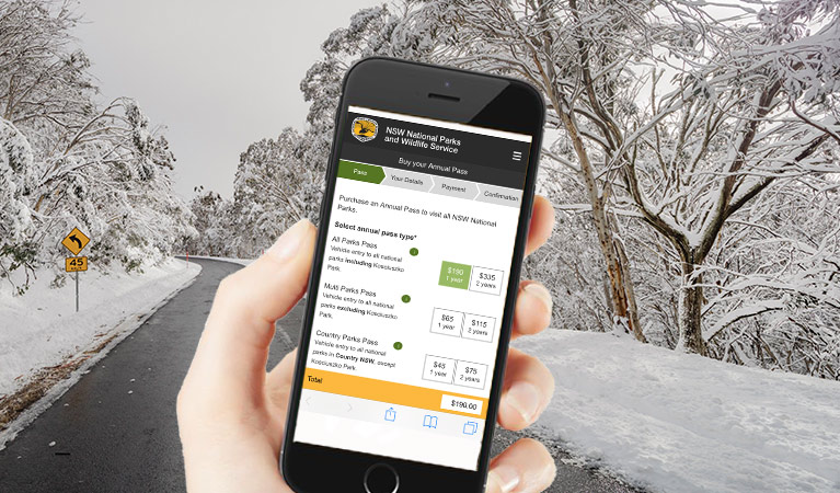A hand holds a mobile phone in front of a snow-covered road in Kosciuszko National Park. Photo: Murray Vanderveer Artwork: DPIE