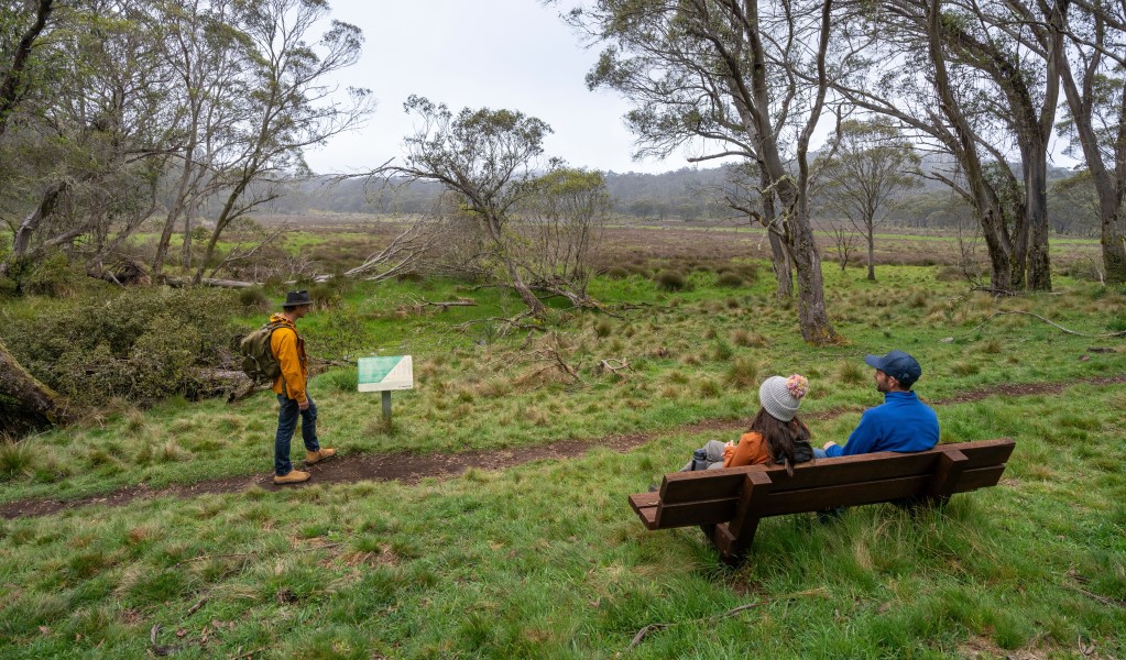 Hikers resting on a wooden bench and reading an interpretive sign on Polblue Swamp track in Barrington Tops National Park. Photo: John Spencer &copy; DCCEEW