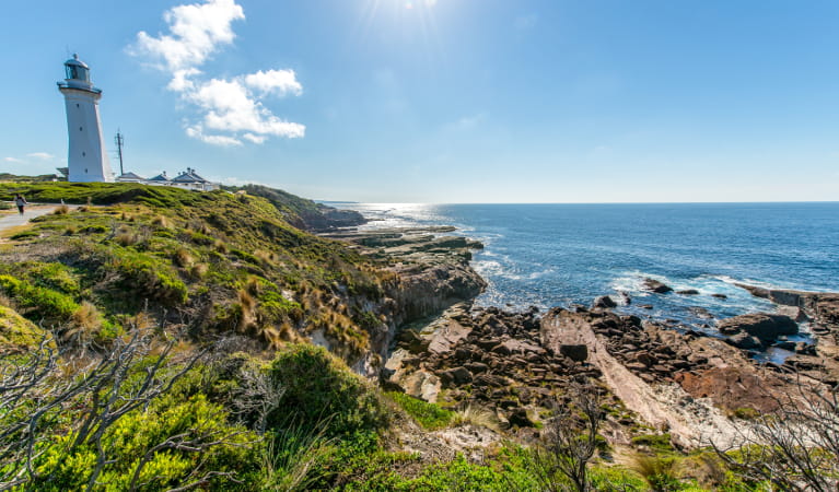Distant view of Green Cape Lighthouse and outbuildings set on rugged coastal terrain with rocky reefs. Photo: John Spencer &copy; DPIE