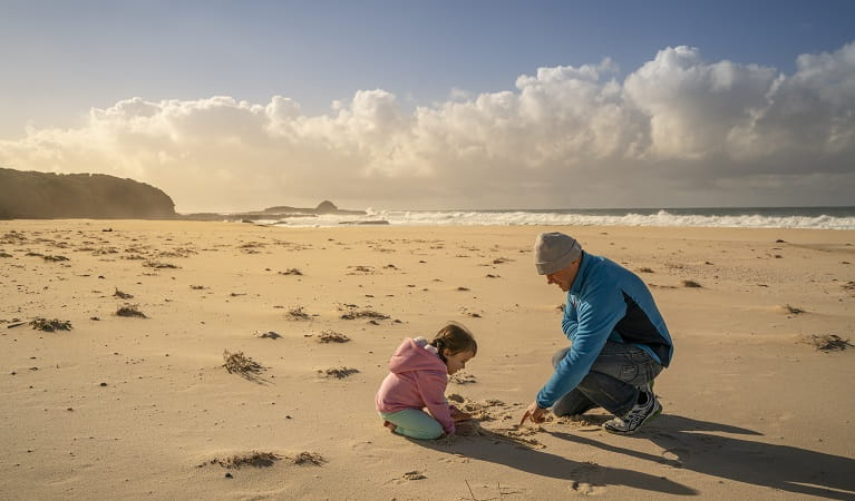 A father and baby daughter playing on the sand at the beach, Haycock Point picnic area, Beowa National Park. Photo: John Spencer/OEH