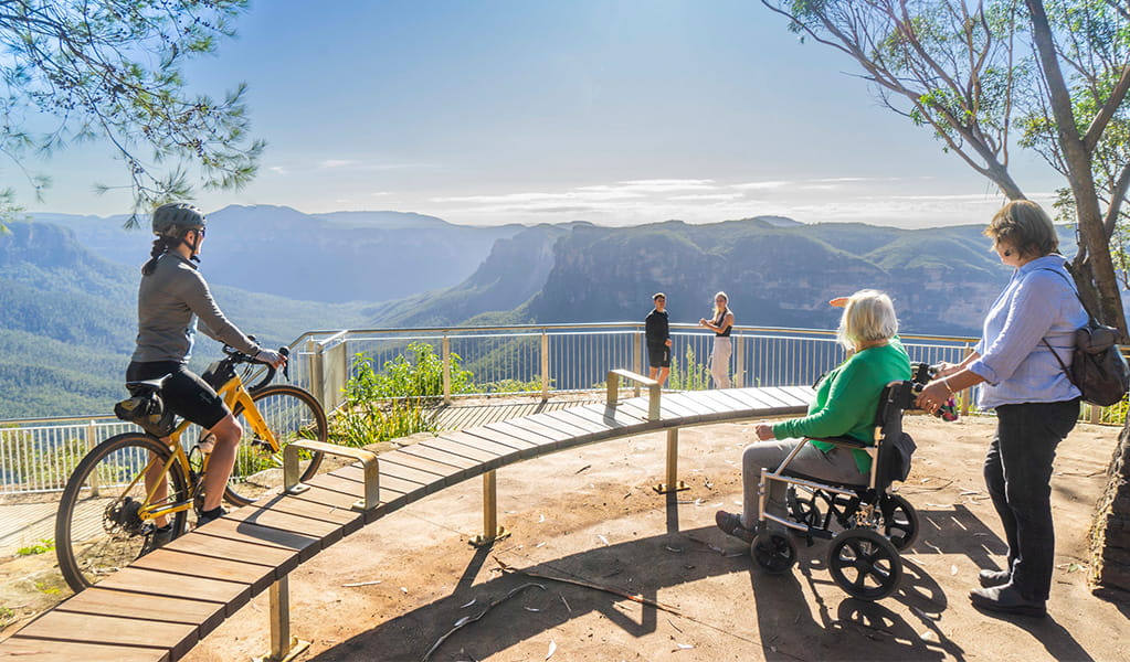 A cyclist on a bike, a person with reduced mobility in a wheelchair and other visitors at Evans lookout in Blue Mountains National Park. Photo credit: Simone Cottrell/DPE &copy; DPE
