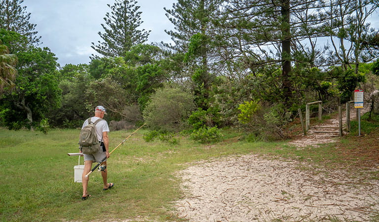 A man with a fishing rod walks across the grass toward a sandy trail and small set of steps. Photo credit: John Spencer &copy; DPIE
