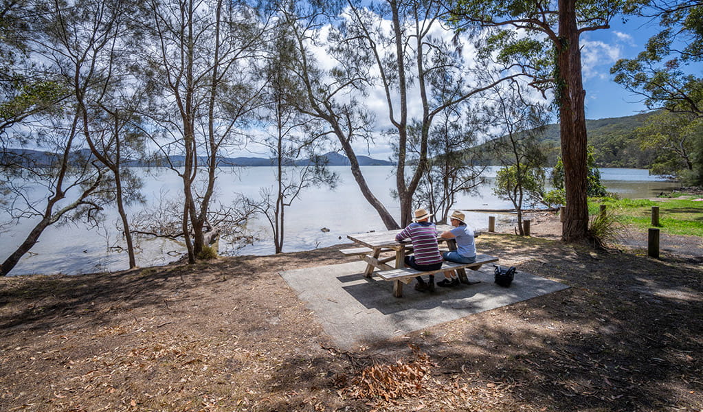 2 people sitting at a picnic table looking at the view of Wallis Lake at Sunset picnic area in Booti Booti National Park. Credit: John Spencer &copy; DPE