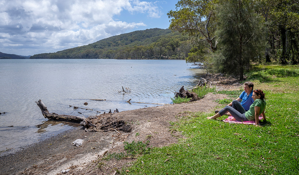 2 people sitting on a picnic rug looking at the view of Wallis Lake at Sunset picnic area in Booti Booti National Park. Credit: John Spencer &copy; DPE