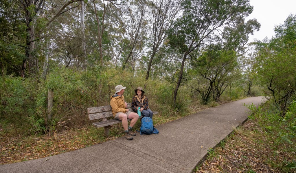 Two people sitting on a bench next to the path that leads to The Lookdown lookout in Bungonia National Park. Photo: John Spencer &copy; DCCEEW