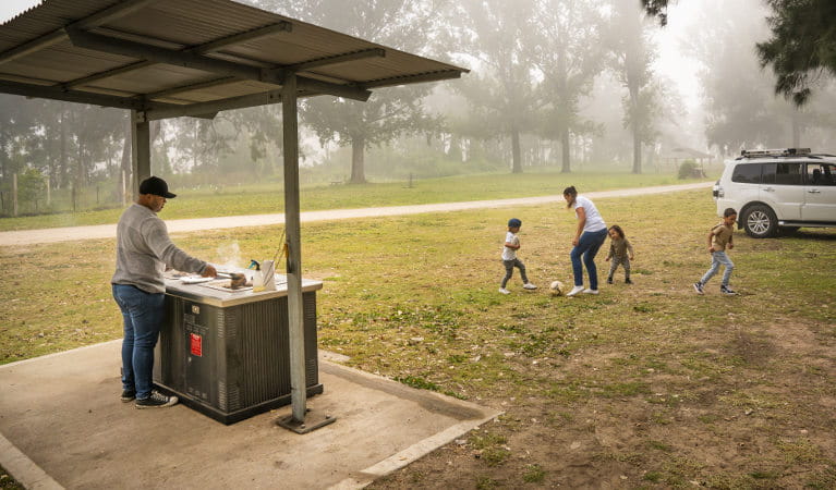 A man cooks up a barbecue lunch as a woman and three kids play soccer in the background. Cattai Farm picnic area in Cattai National Park. Photo: John Spencer/OEH