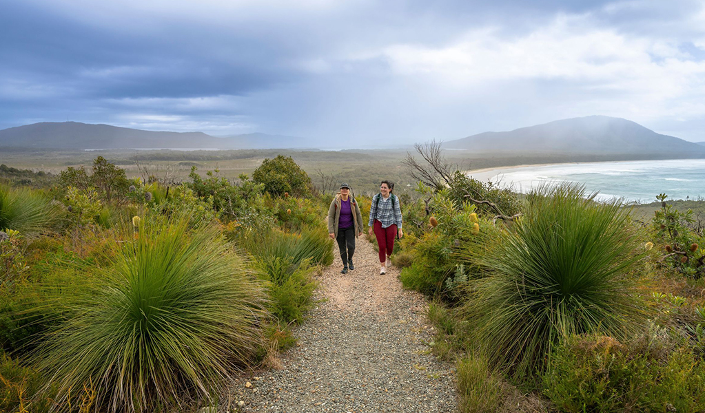 2 people on a track along Diamond Head loop walk surrounded by coastal heath with Dunbogan beach and Perpendicular Point in the background. Credit: John Spencer &copy; DCCEEW