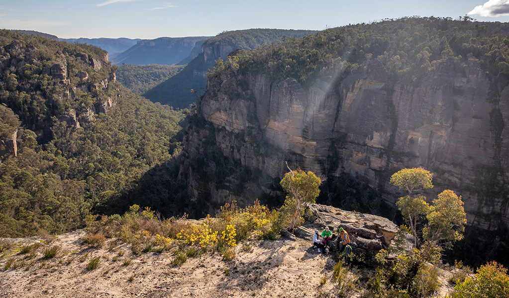 An aerial view of 3 hikers at Carne View lookout along Broad Swamp to Birds Rock walking track with sheer cliffs above Carne Creek in the background. Credit: John Spencer &copy; DCCEEW