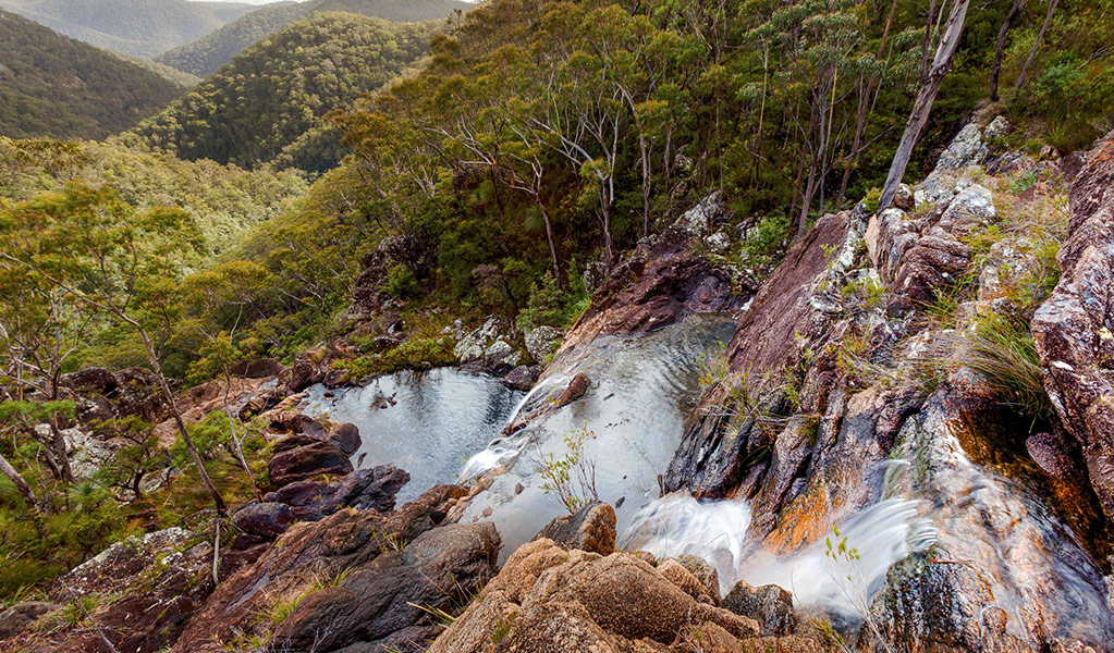 View from above of falls cascading down multiple rock ledges in a rugged wilderness setting of ridges and valleys. Photo credit: Robert Cleary &copy; DPIE