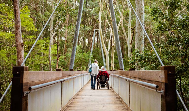 A woman and a man in a wheelchair cross a bridge on Yuelarbah walking track, Glenrock State Conservation Area. The bridge is made of wood and metal, and surrounded by trees. Photo: Jared Lyons &copy; DPE