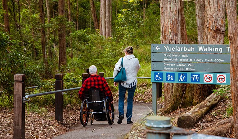 A man in a wheelchair and a woman explore Yuelarbah walking track in Glenrock State Conservation Area. The track is sealed and level and weaves through forest with tall trees on all sides. Photo: Jared Lyons &copy; DPE