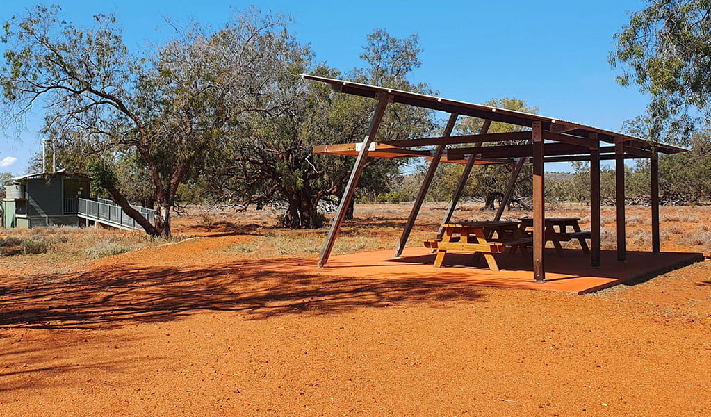 View of a picnic table with a shelter near toilet facilities, set on red desert earth amongst scattered trees. Photo credit: Jessica Ellis &copy; DPIE