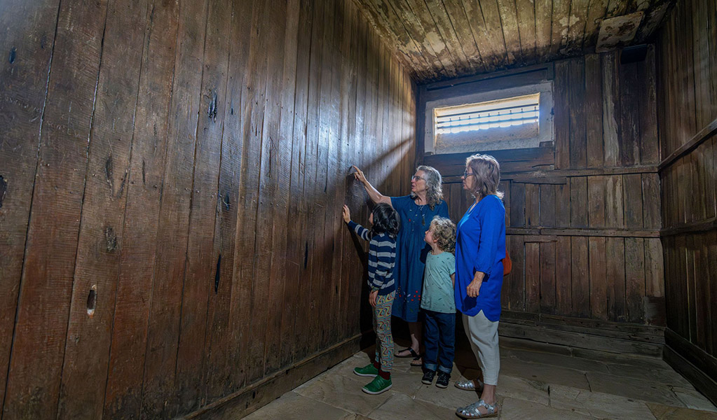 4 visitors inside an old jail cell looking at graffiti left by convicts on the cell walls inside Hartley Courthouse. Credit: John Spencer &copy; DCCEEW