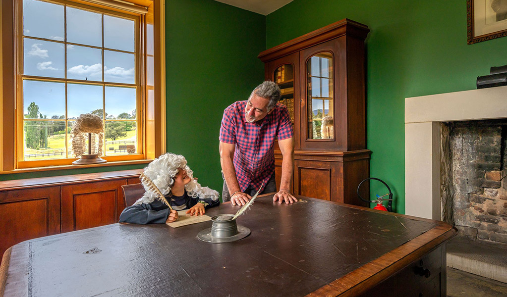 A child dressed as a judge pretending to write with a feather quill with an adult at a desk inside Hartley Courthouse. Credit: John Spencer &copy; DCCEEW