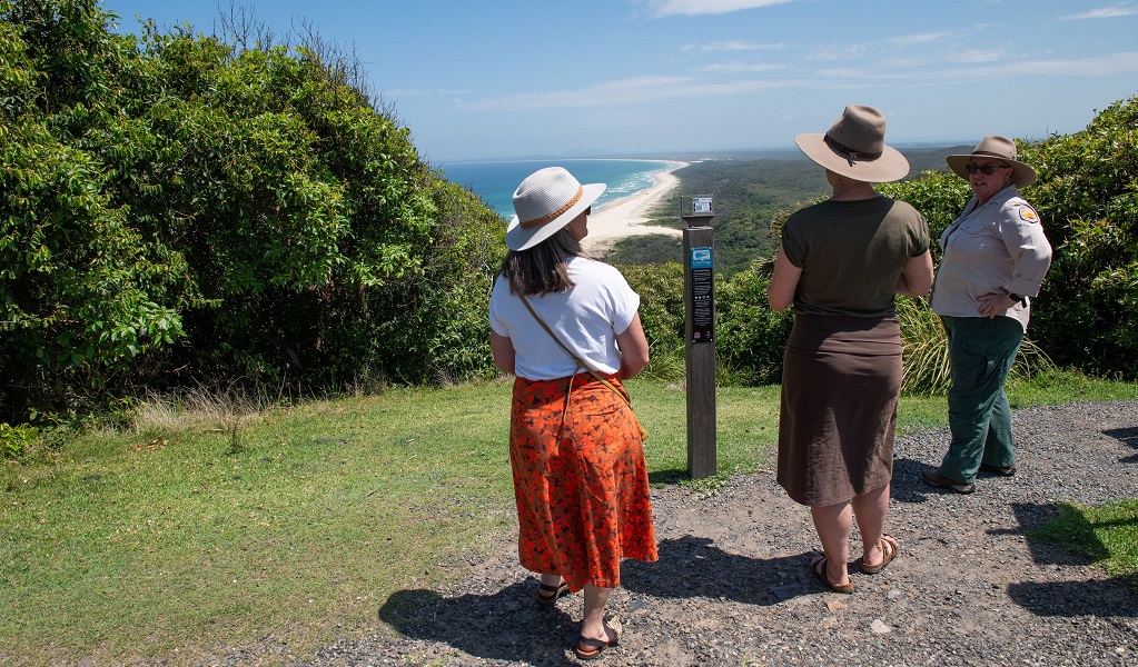 Visitors and NPWS guide chat on the lighthouse lawn, enjoying the view of white sand beaches, lush greenery and the clear blue ocean, Smoky Cape Lighthouse, Hat Head National Park. Photo: Louise Walpole/DCCEEW &copy; DCCEEW
