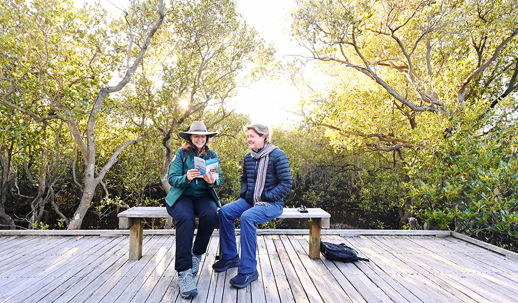 A ranger and a visitor looking at a book about shorebirds while resting on a bench surrounded by mangroves. Credit: Adam Hollingworth &copy; DCCEEW