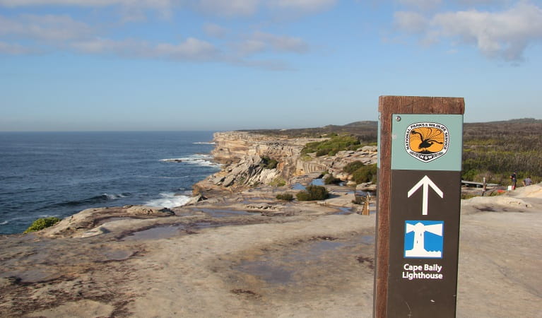 Park sign pointing to Cape Baily lighthouse, set against a vista of ocean, rugged rocky coastline and bushland.  Photo: Natasha Webb/DPIE