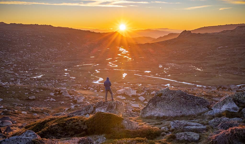 A hiker watching the sun set over the mountains at Muellers Pass in Kosciuszko National Park. Credit: Jan Breckwoldt &copy; DCCEEW