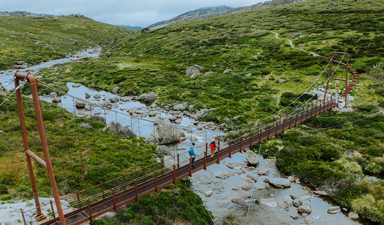 An aerial image of 2 people crossing Spencers Creek suspension bridge with a flowing creek and alpine landscape in the background. Credit: Daniel Parsons / DCCEEW &copy; Caravel Content
