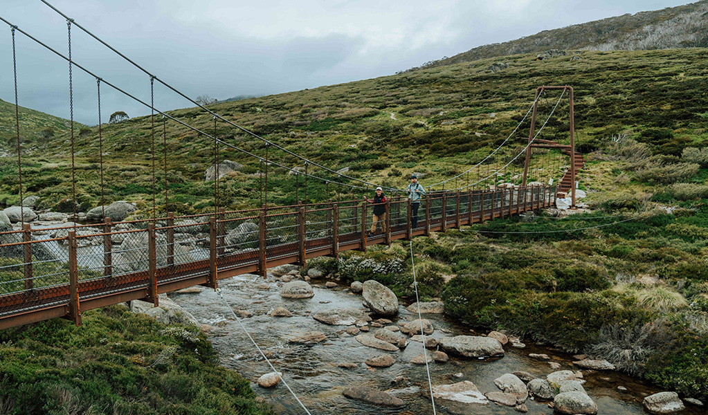 2 people crossing Spencers Creek suspension bridge with a flowing creek and alpine landscape in the background. Credit: Remy Brand / DCCEEW © Caravel Content