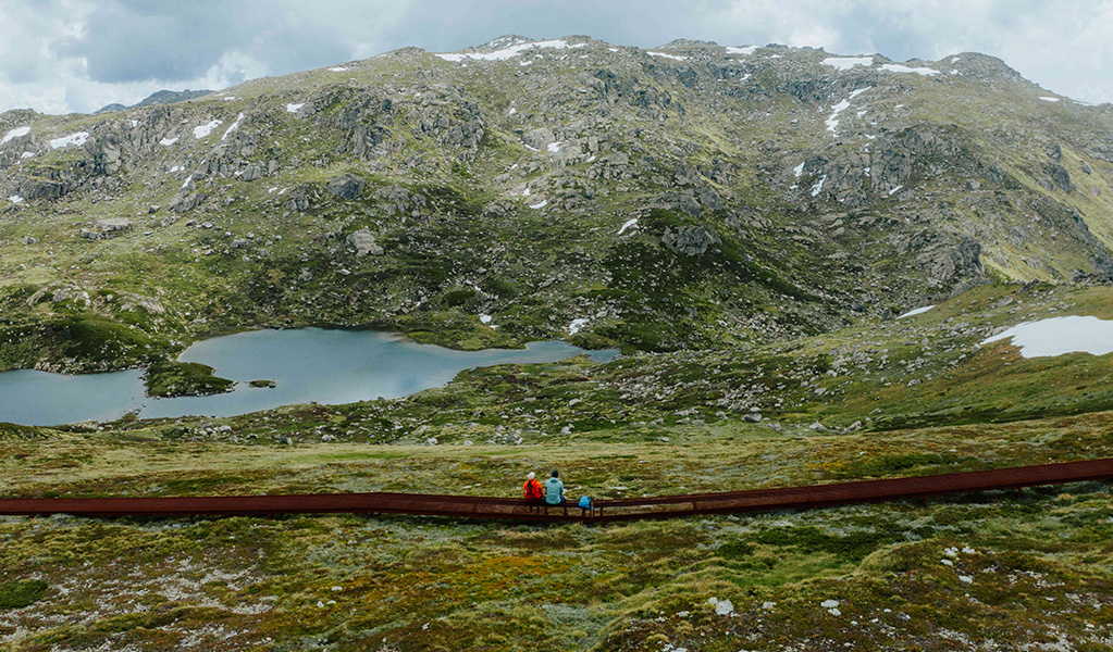 2 hikers on a raised path admiring snow capped Main Range mountain views on day 2 of Snowies Alpine Walk. Credit: Daniel Parsons / DCCEEW © Caravel Content