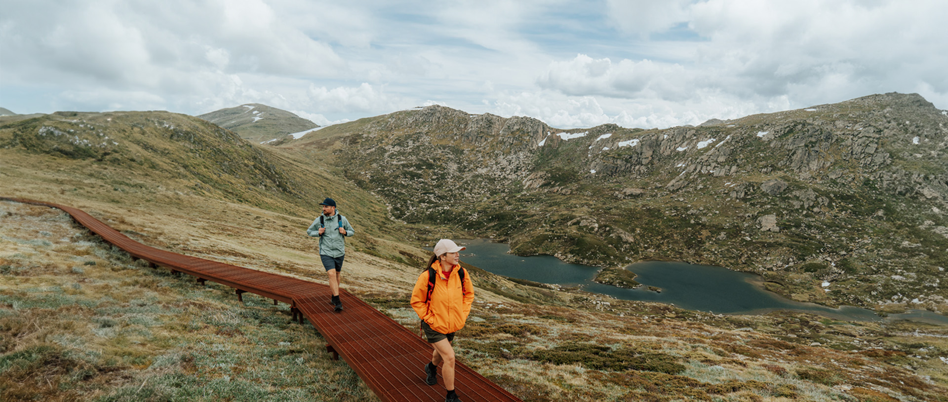 2 hikers walking on a raised path on day 2 of Snowies Alpine Walk with snow capped mountains in the background. Credit: Remy Brand / DCCEEW © Caravel Content