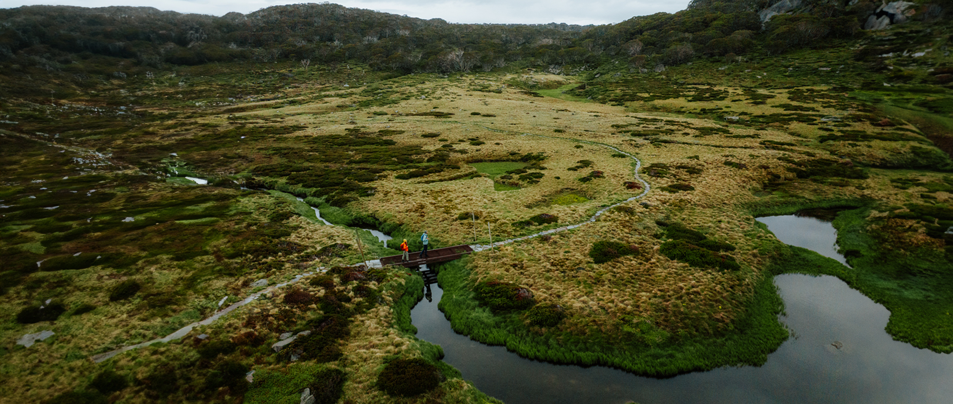 An aerial view of 2 hikers crossing a winding creek surrounded by marsh on Snowies Alpine Walk, Kosciuszko National Park. Credit: Daniel Parsons / DCCEEW © Caravel Content