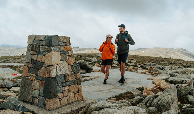 A man and woman walking towards the stone trig at the summit of Mount Kosciuszko with mountain peaks in the background. Credit: Remy Brand / DCCEEW © Caravel Content