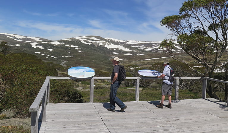 2 bushwalkers read park information at Charlotte Pass lookout, with mountains in the background. Photo: Elinor Sheargold/DPIE.