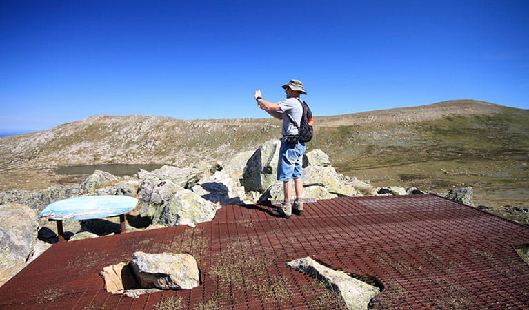 A man takes a photo at Cootapatamba lookout, with Mount Kosciuszko in the background, Kosciuszko National Park. Photo: Elinor Sheargold &copy; DPIE