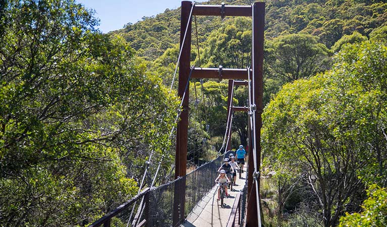 A family of mountain bike riders cycle across a suspension bridge along Thredbo Valley track, Kosciuszko National Park. Photo: Thredbo Resort