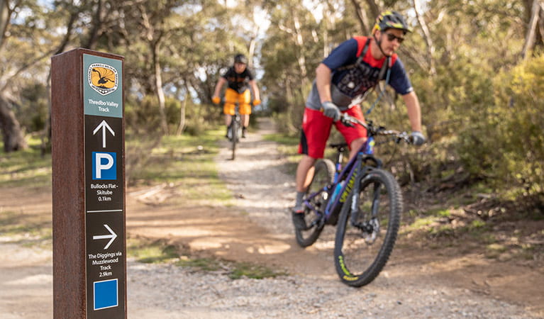 Two mountain bike riders cycle past a sign at the junction of Muzzlewood and Thredbo Valley tracks, Kosciuszko National Park. Photo: Robert Mulally/DPIE