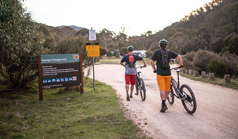 Two mountain bike riders walk past a sign entering Ngarigo campground, Kosciuszko National Park. Photo: Robert Mulally/DPIE