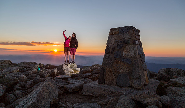 A woman and man take a selfie photo at the summit of Mount Kosciuszko at sunset, Kosciuszko National Park. Photo: Tourism Snowy Mountains