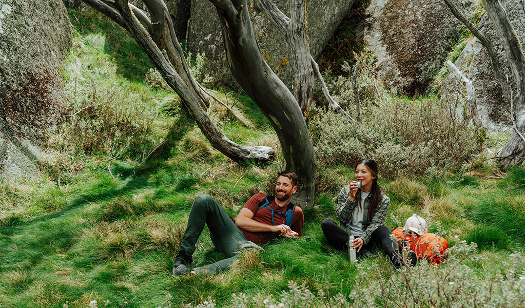 A man and woman sitting on grass enjoying a break surrounded by snow gums and large boulders along Perisher Valley to Bullocks Flat walk. Credit: Remy Brand / DCCEEW &copy; Caravel Content