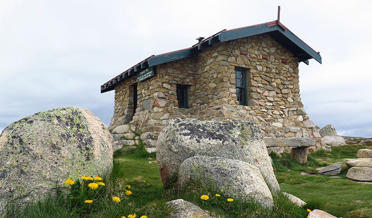 View of historic stone hut set in alpine grassland, with large boulders and wildflowers in the foreground. Photo: Stephen Townsend &copy; DPIE