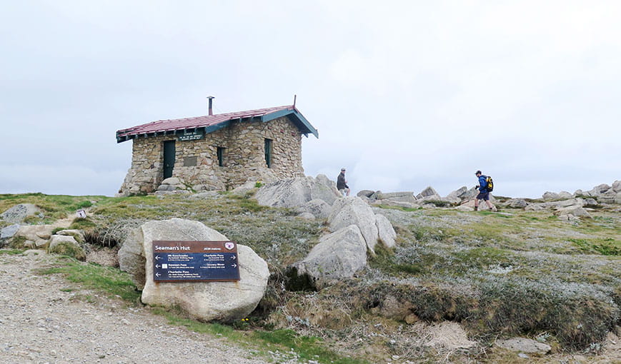 View of hikers passing historic Seamans Hut, with gravel trail and large boulders in the foreground. Photo: Stephen Townsend &copy; DPIE