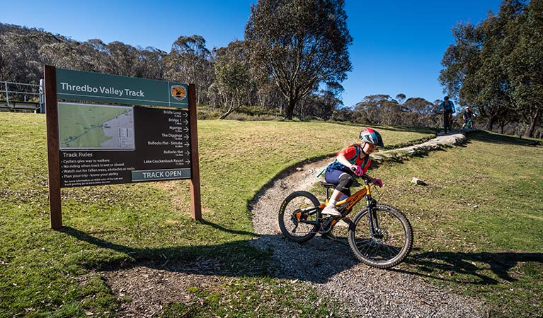 A young girl mountain bikes past a sign at the start of Thredbo Valley track, Kosciuszko National Park. Photo: Robert Mulally/DPIE