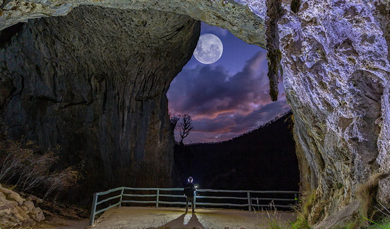 A visitor stands inside the entrance to North Glory Cave, looking out towards Kosciuszko National Park at dusk with a full moon in the sky. Photo: Adam Klumper &copy; Adam Klumper